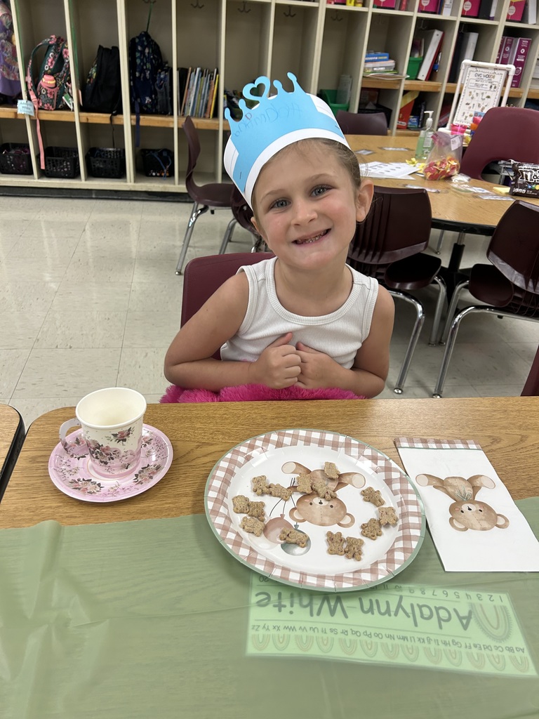 A kindergarten student wearing a blue crown smiles while sitting at a decorated table with a plate of teddy bear cookies and a pink teacup.