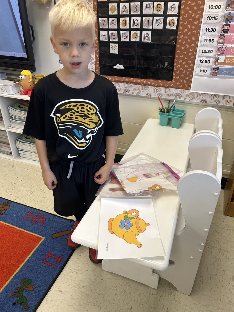 A kindergarten student wearing a Jacksonville Jaguars shirt stands by a desk with a large paper teapot cutout.