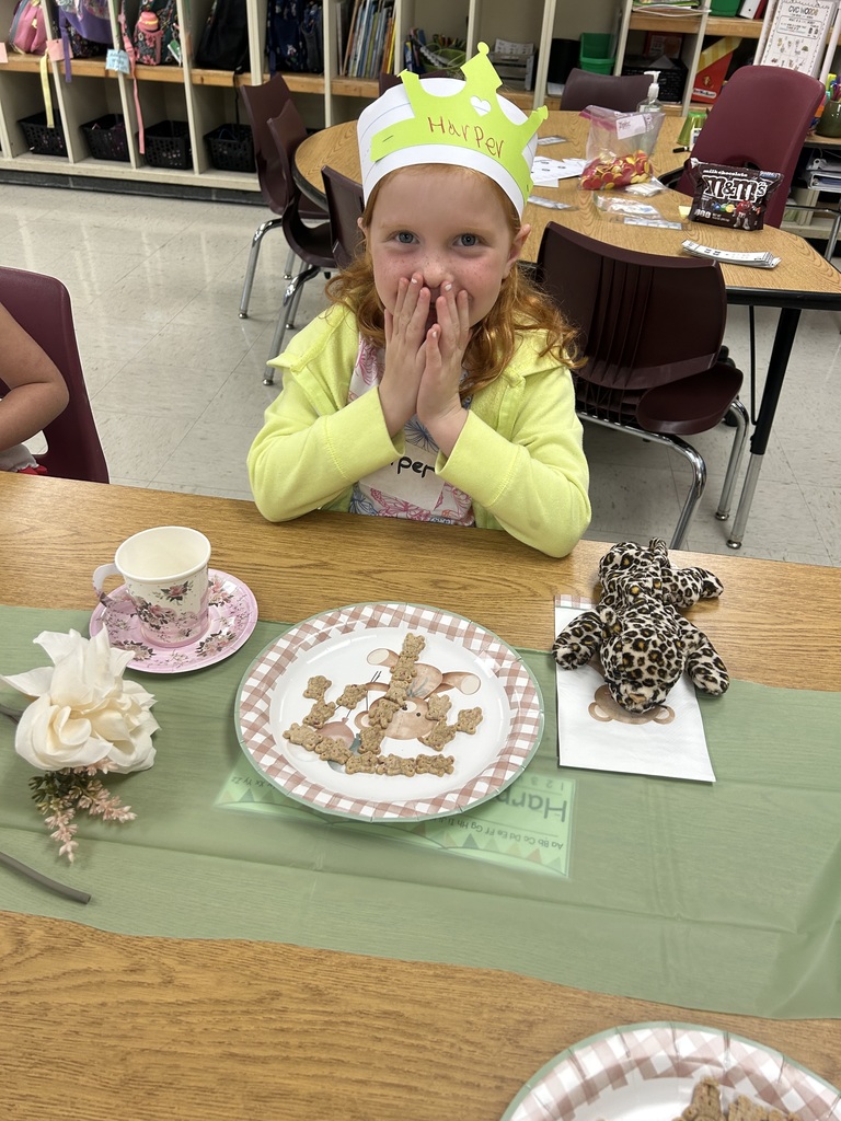 A kindergarten student wearing a paper crown labeled “Harper” smiles with hands over her mouth while sitting at a table set with teddy bear–themed plates, cookies, and a teacup.