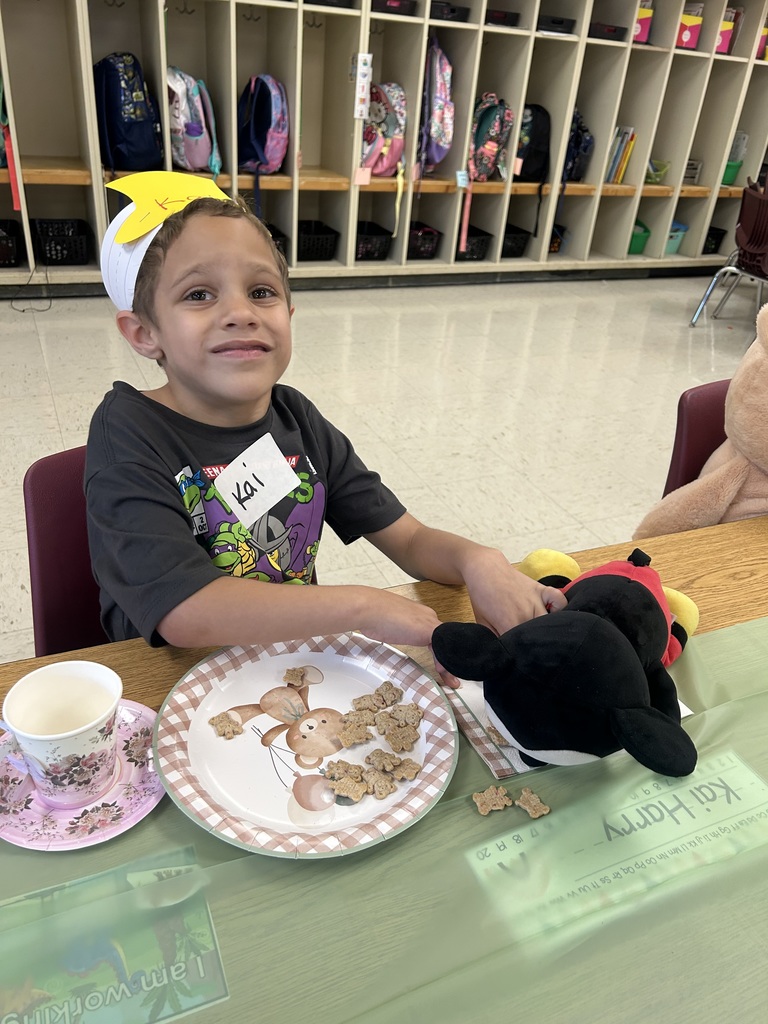 A kindergarten student wearing a yellow crown sits at a decorated table with a plate of teddy bear cookies, a teacup, and a teddy bear napkin.