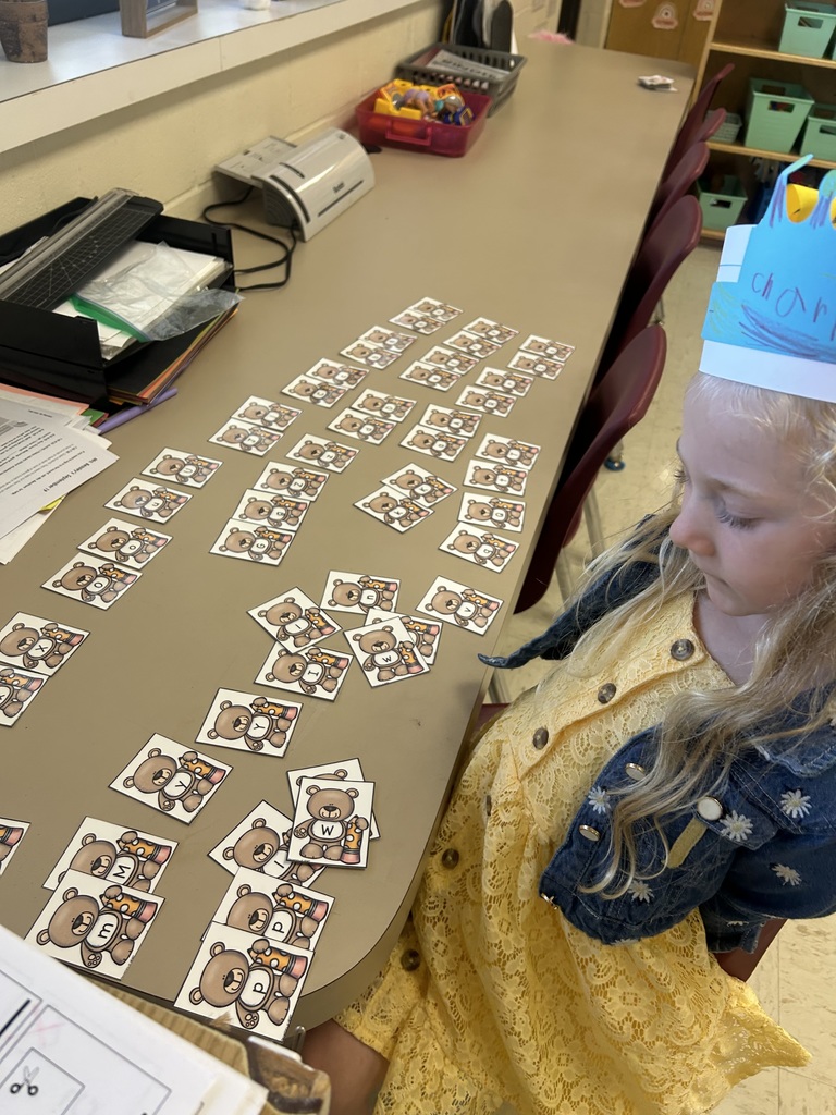 A kindergarten student in a yellow dress and denim jacket sits at a table arranging teddy bear–themed letter cards.
