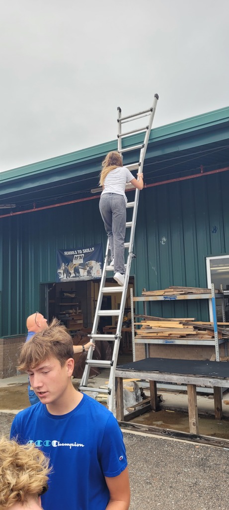 A Yulee Middle School student climbs a ladder outside a green metal building while a Yulee High School construction teacher supervises and classmates watch, learning about ladder safety.