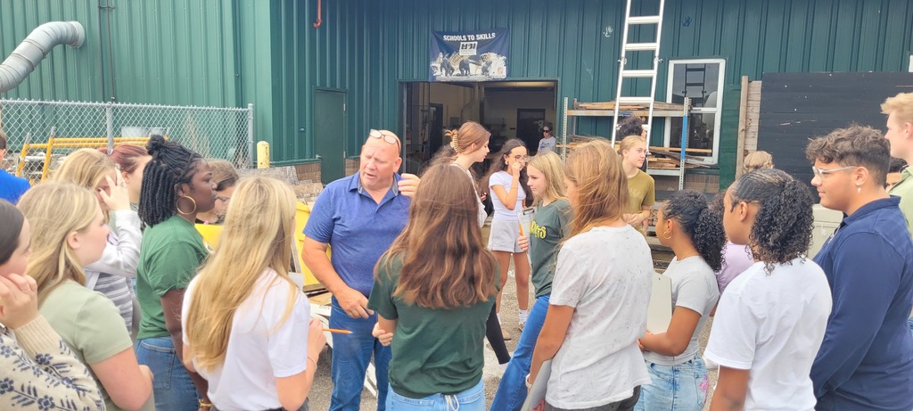 A group of students gather around the Yulee High School construction teacher outside a green metal workshop, listening to an explanation about ladder safety and class projects.