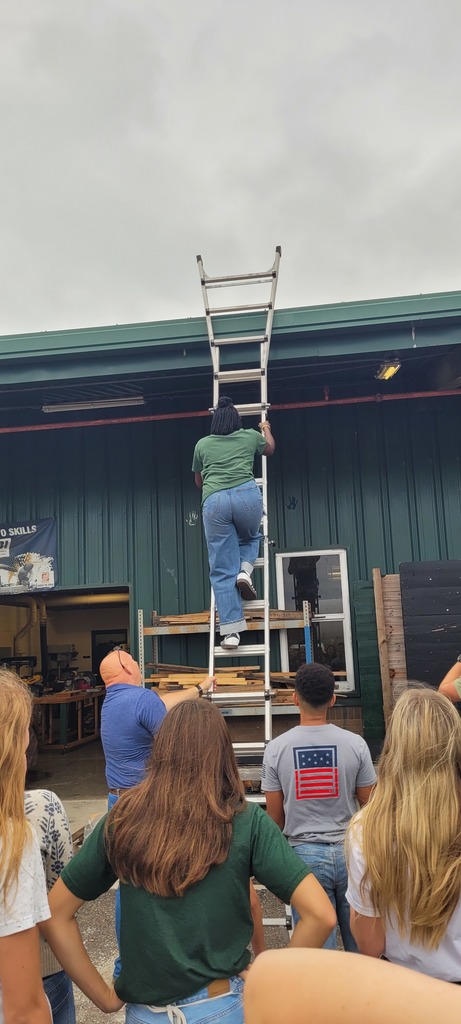 A Yulee Middle School student climbs a ladder outside a green metal building while a Yulee High School construction teacher supervises and classmates watch, learning about ladder safety.