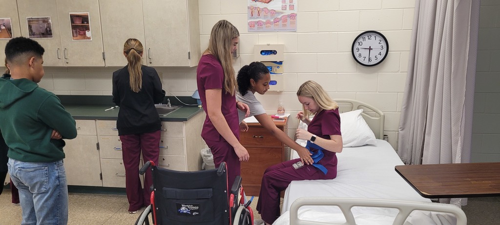 High school CNA students demonstrate safe patient transfer techniques using a hospital bed setup, while visiting middle school students observe the process.