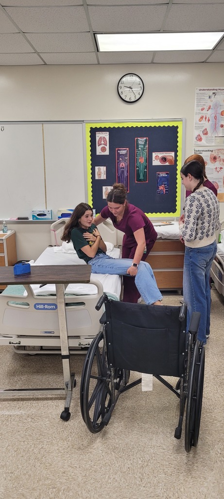 A high school CNA student in maroon scrubs assists a visiting Yulee Middle School student in safely transferring from a hospital bed during a classroom demonstration. A wheelchair and anatomy posters are visible in the background.