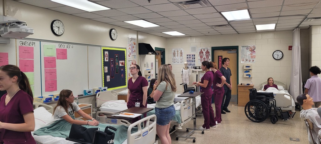 A Yulee High School CNA classroom filled with students practicing patient care techniques such as bed transfers and vital sign monitoring, with anatomy charts and medical equipment visible.