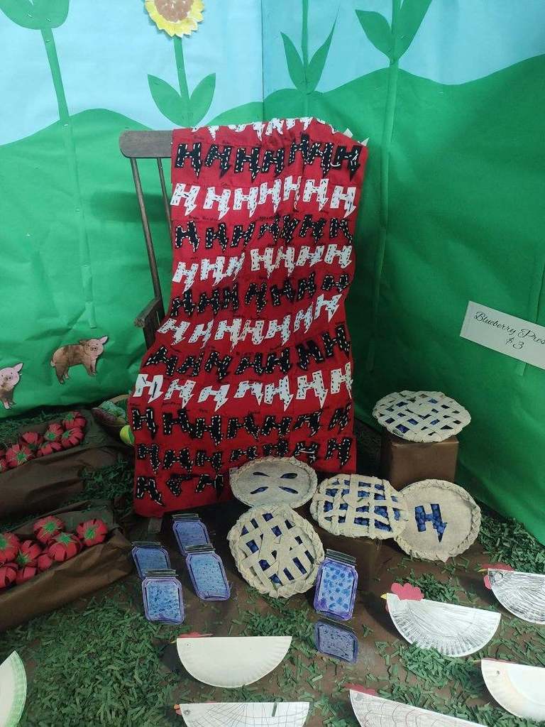 A red quilt with white and black “H” letters draped over a chair beside handmade paper pies, jars, and chickens arranged on green grass-like paper and surrounded by sunflower decorations.