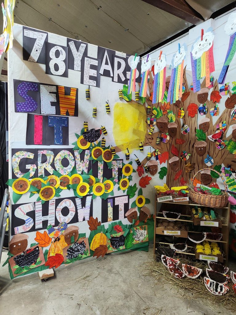 A display with large cutout letters reading “78 Years Sew It Grow It Show It,” surrounded by handmade paper bees, sunflowers, acorns, and colorful rainbow crafts, celebrating a farm-themed art showcase.