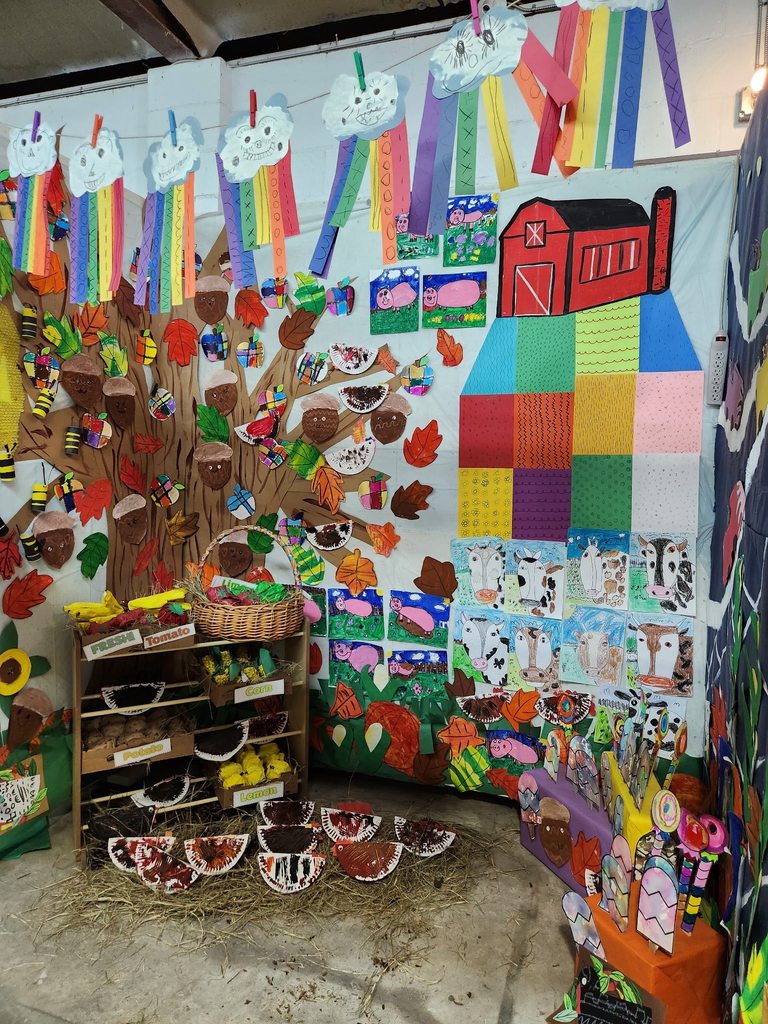 A cozy barn-themed booth featuring hay on the floor, small scarecrow figures, string lights, and children’s artwork of fall leaves, trees, and farm animals displayed on black walls.