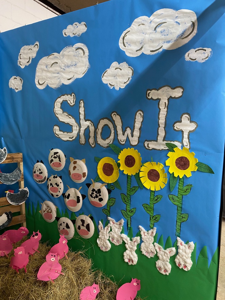 A light blue wall with clouds, sunflowers, paper cows, bunnies, and pink pigs made from paper plates and cotton balls. The text “Show It” is displayed above, tying into the fair’s theme.