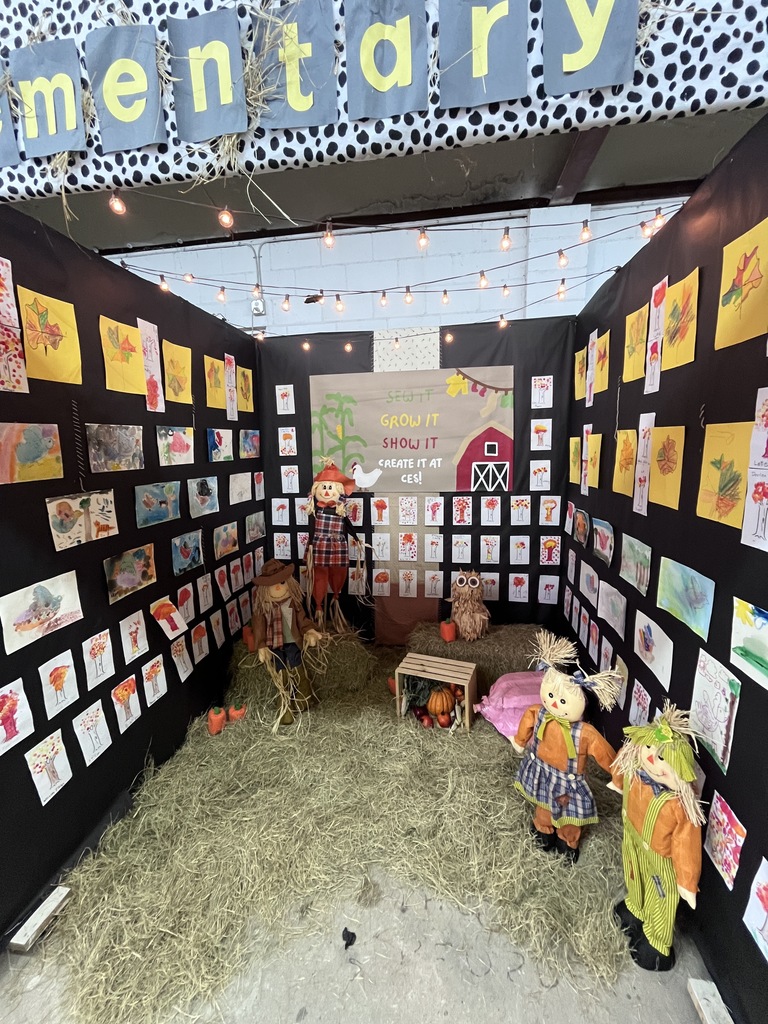 A cozy barn-themed booth featuring hay on the floor, small scarecrow figures, string lights, and children’s artwork of fall leaves, trees, and farm animals displayed on black walls.
