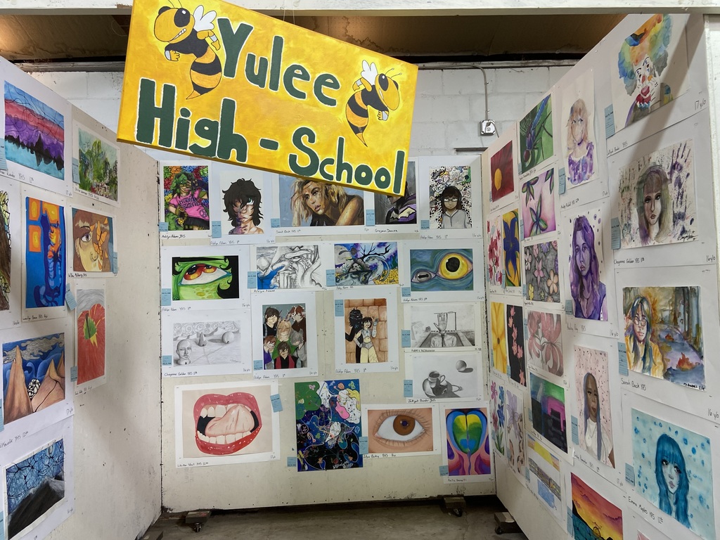 A yellow banner reading “Yulee High School” with cartoon hornets hangs above a display of colorful student artwork, including portraits, surrealist paintings, and sketches arranged across three walls.