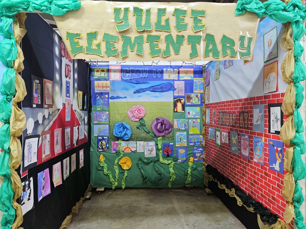 A booth decorated with large green and gold paper vines and a burlap sign reading “Yulee Elementary.” The walls are covered with bright student artwork featuring flowers, skies, and animals surrounding a “Grow It” theme.