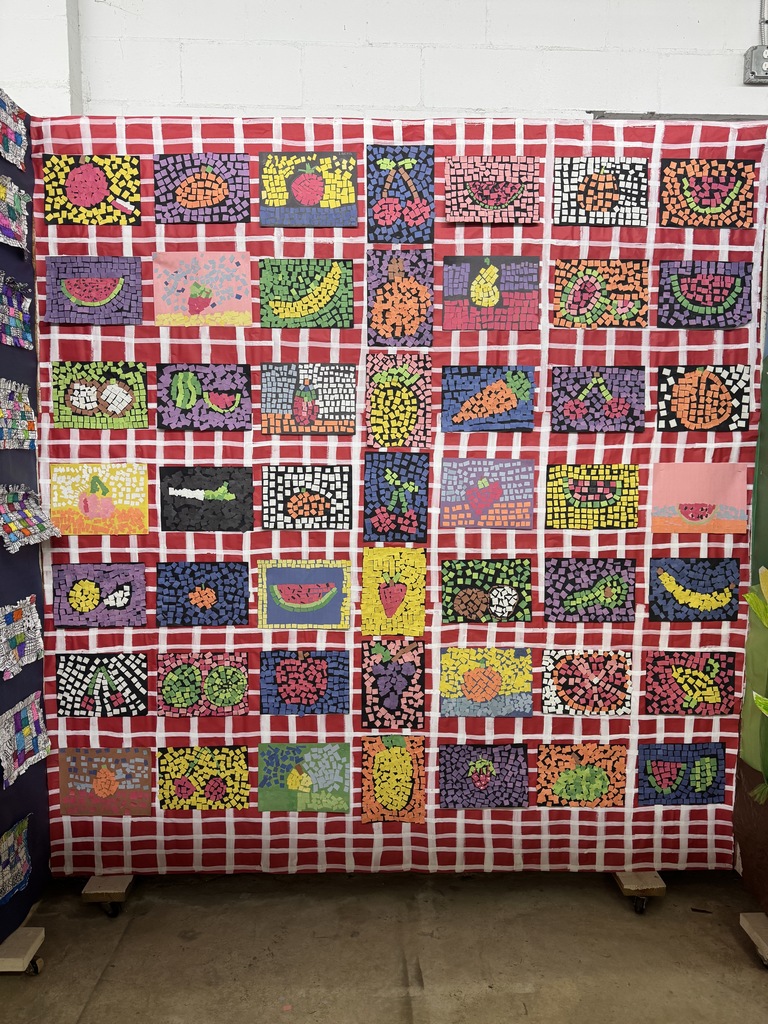 A wall covered with colorful mosaic fruit artworks, each framed against a red-and-white checkered picnic blanket background, showcasing fruits like watermelon, bananas, and berries.
