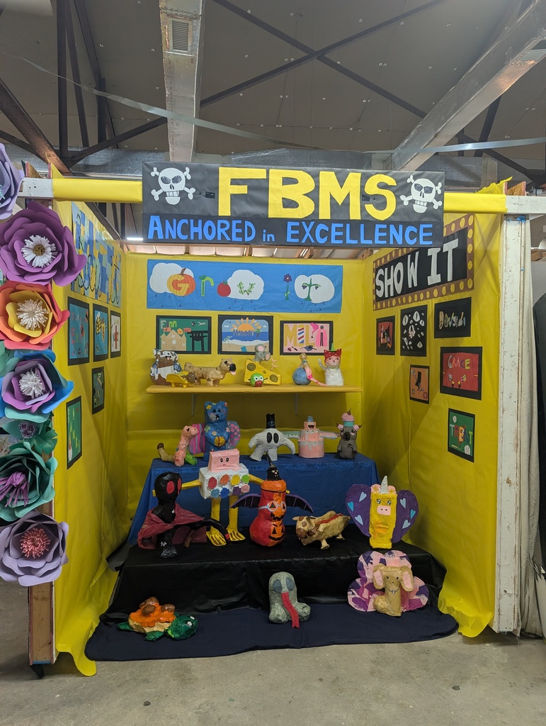 A yellow and black art booth with a banner reading “FBMS Anchored in Excellence.” The display features large paper flowers and shelves filled with colorful clay and papier-mâché sculptures made by students.