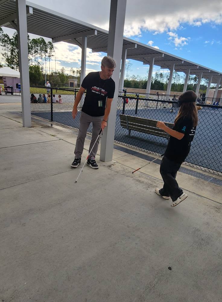 An adult instructor and a student wearing a blindfold practice mobility techniques using white canes under a covered walkway at school.