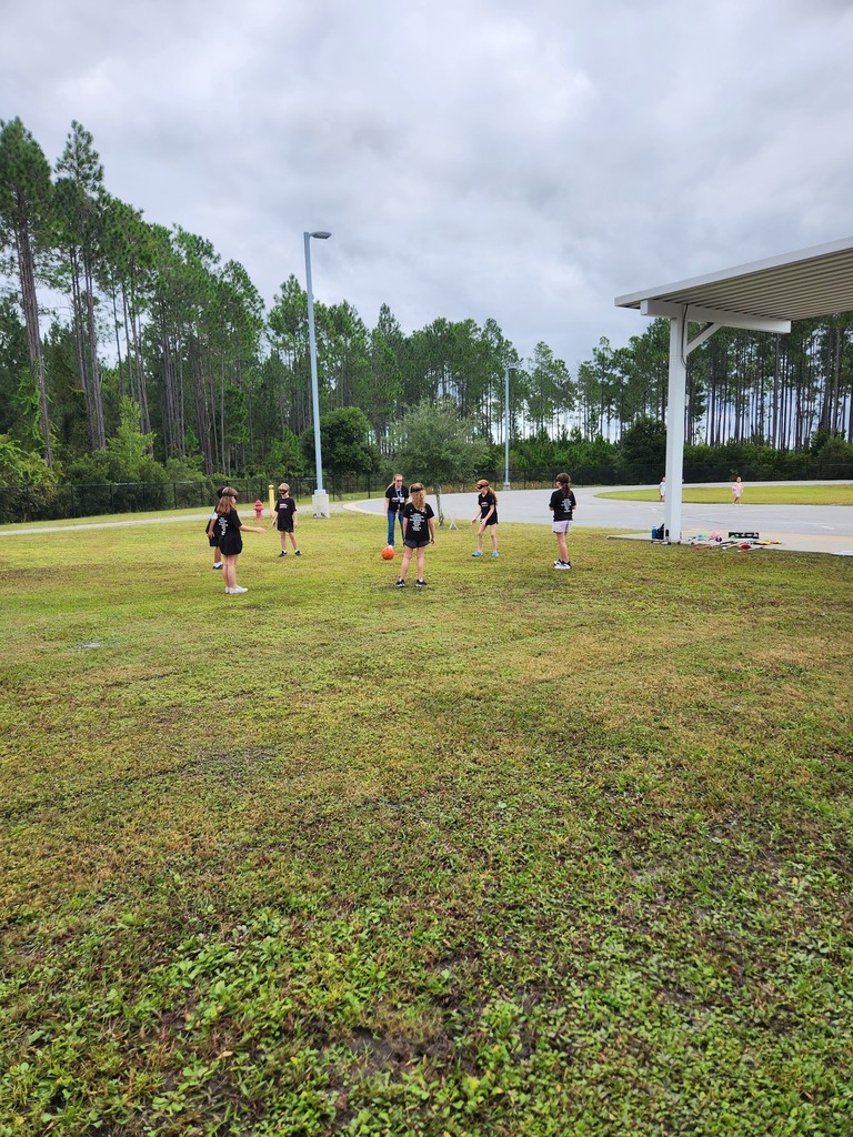 Students wearing black shirts and blindfolds stand on a grassy field playing a game with a bright orange ball during an outdoor White Cane Day activity.