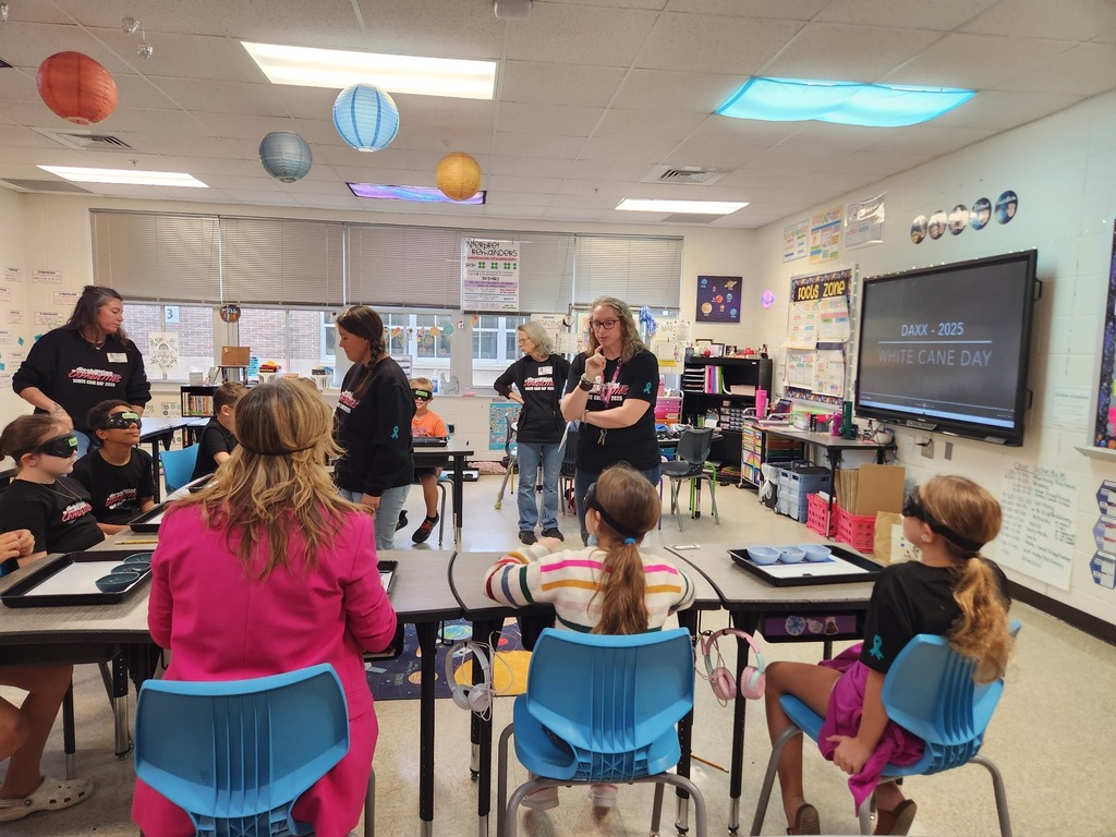 Students wearing blindfolds sit at tables while teachers guide them through activities for White Cane Day. A presentation slide on the board reads “DAXX – 2025 White Cane Day.”