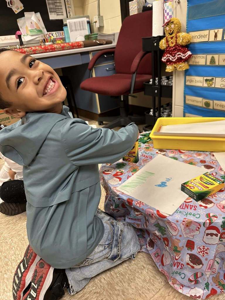A kindergarten student smiles while participating in a winter-themed classroom activity involving drawing and hands-on learning materials at a small table.
