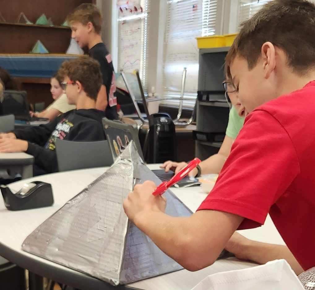 A sixth-grade student uses a marker to label a model pyramid constructed from cardboard while classmates work at desks in the background.