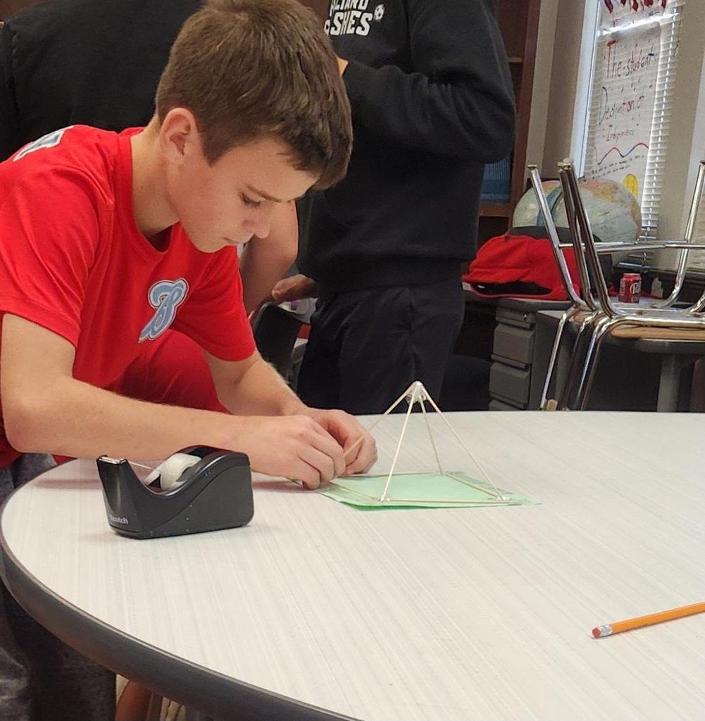 A sixth-grade student assembles a small pyramid structure made of sticks and paper at a classroom table, focusing on construction details.
