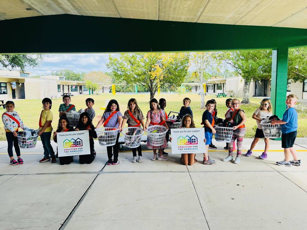 A group of Yulee Primary School Safety Patrol students stands in a school walkway holding laundry baskets filled with donated socks, with two students holding signs for the Coalition for the Homeless of Nassau County.