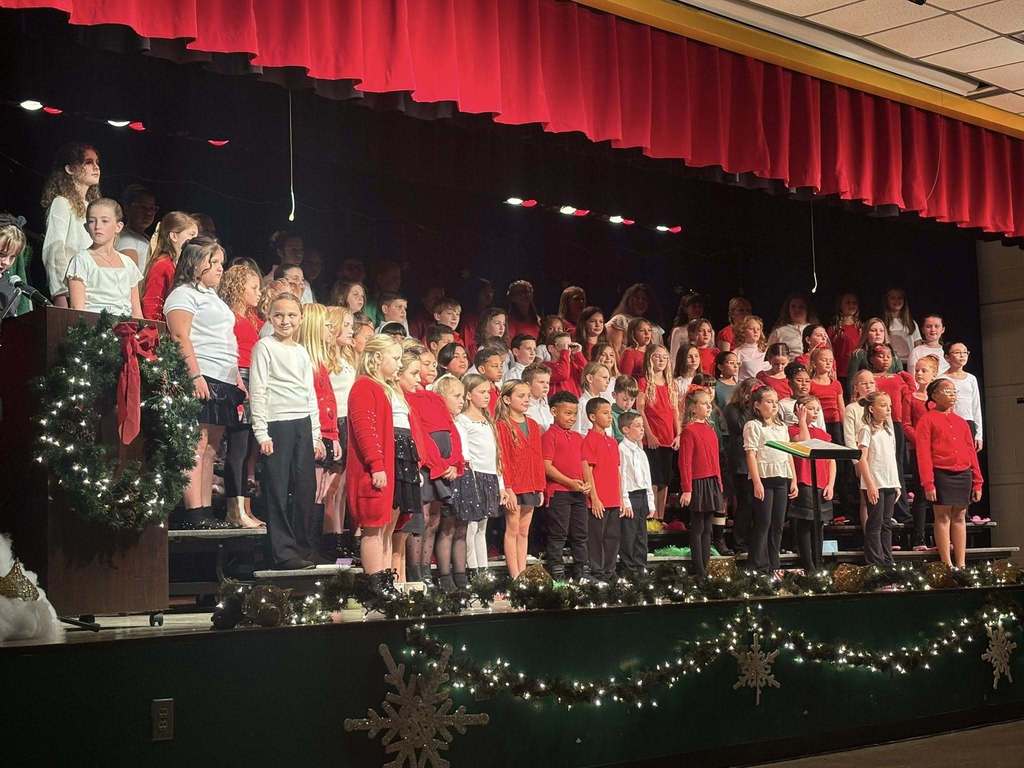 Members of the Yulee Elementary Hornet Singers Choir stand on a decorated school stage wearing red, white, and black attire during a holiday concert performance.