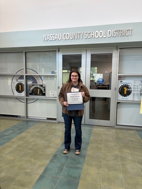An adult education student stands outside the Nassau County School District building holding a certificate of completion.
