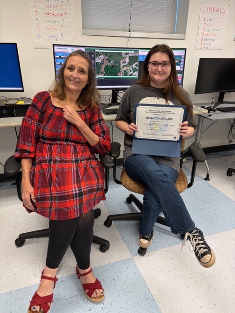 An adult education student poses with an instructor in a classroom setting while holding a certificate from the 911 Telecommunications Dispatch Integrated Education and Training Program.
