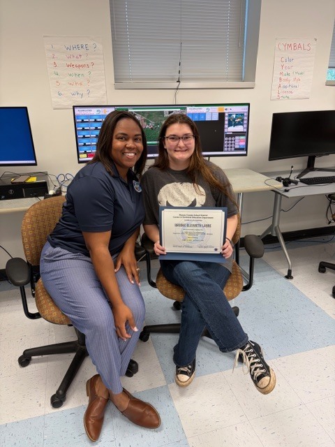 An adult education student poses with an instructor in a classroom setting while holding a certificate from the 911 Telecommunications Dispatch Integrated Education and Training Program.