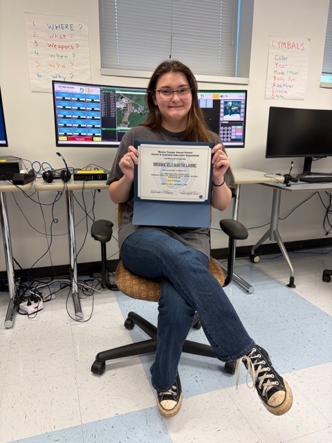 An adult education student sits in a classroom holding a 911 Telecommunications Dispatch program certificate, with computer monitors and training equipment visible behind her.