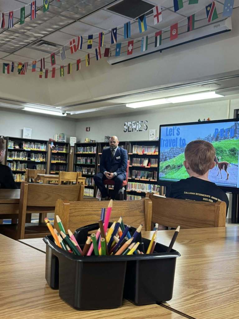 Florida’s Teacher of the Year sits in the Callahan Intermediate School library speaking with students seated at tables, with bookshelves and classroom materials surrounding the group.