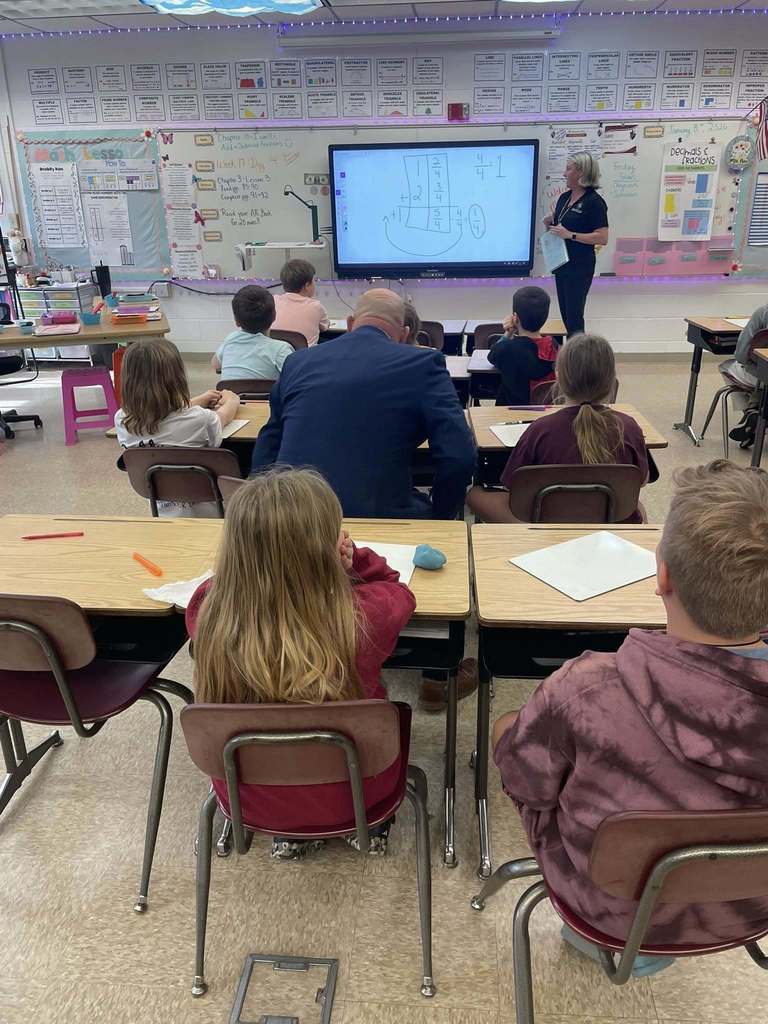 Students at Callahan Intermediate School sit at their desks facing an interactive display as a teacher explains a math lesson, with a visiting educator seated among the students.