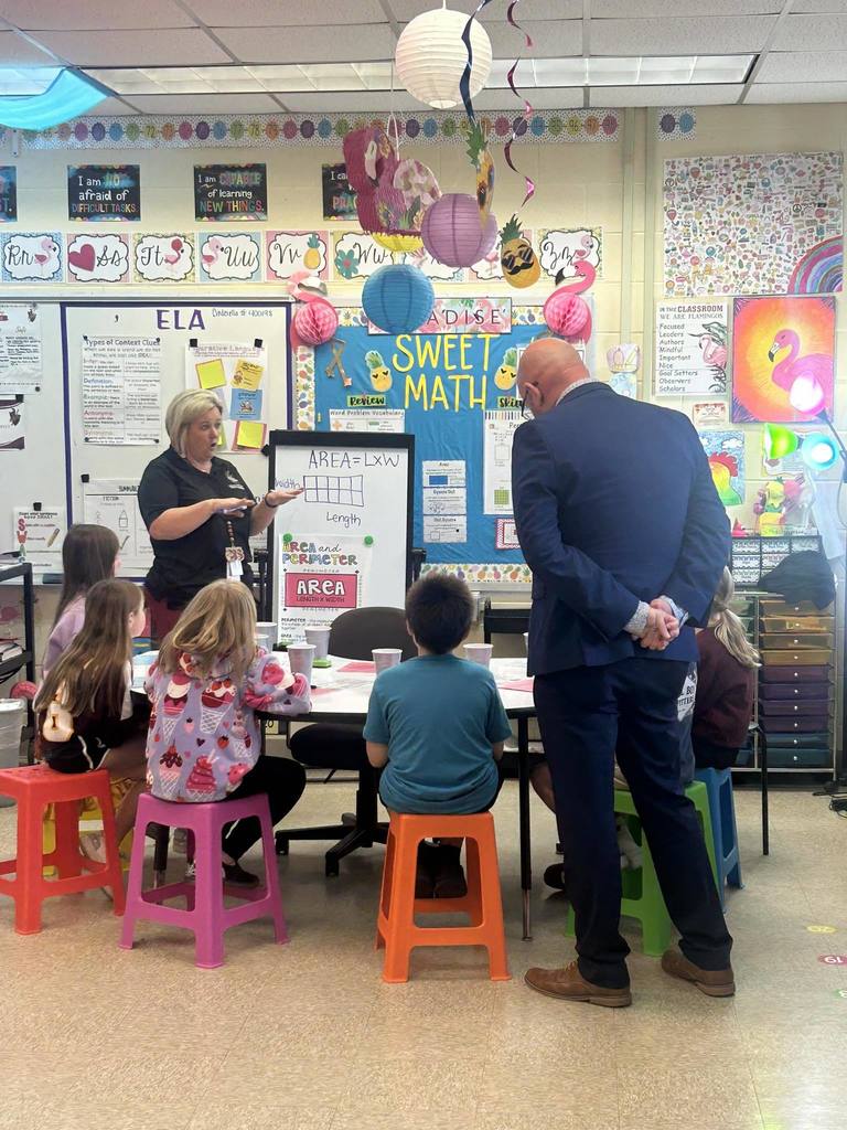 A teacher leads a small-group math lesson at Callahan Intermediate School as students sit on colorful stools around a table, while a visiting educator observes from behind.