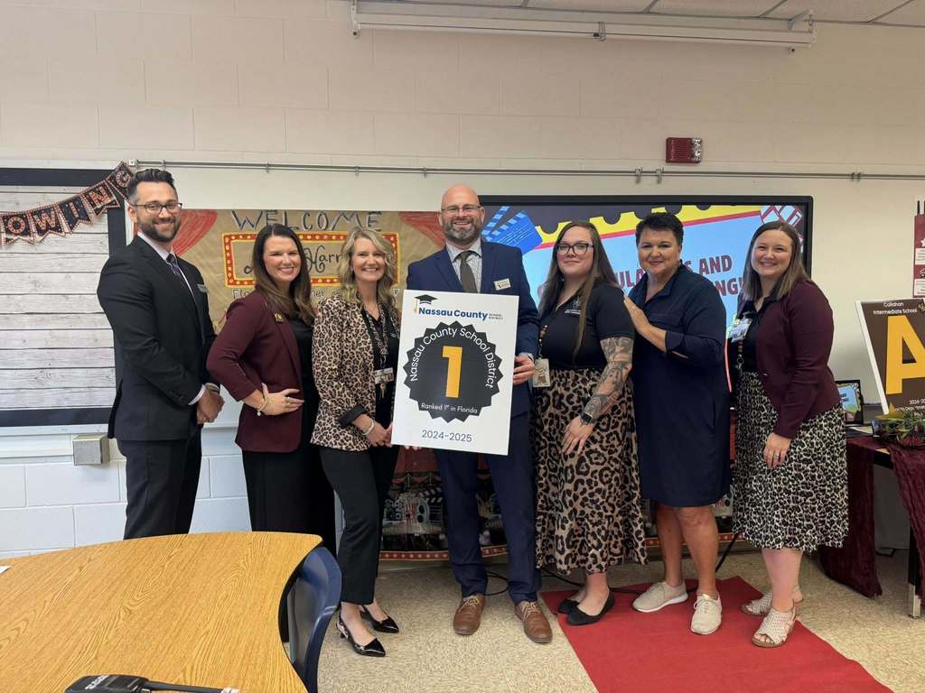 School and district staff stand together inside a classroom at Callahan Intermediate School holding a recognition sign, smiling as they mark the visit from Florida’s Teacher of the Year.