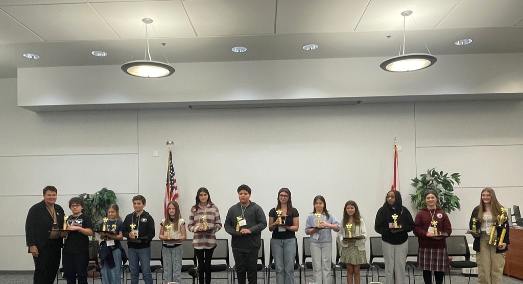 Spelling bee participants stand in a line on stage holding trophies during the Nassau County School District Spelling Bee, with the American and Florida flags displayed behind them.