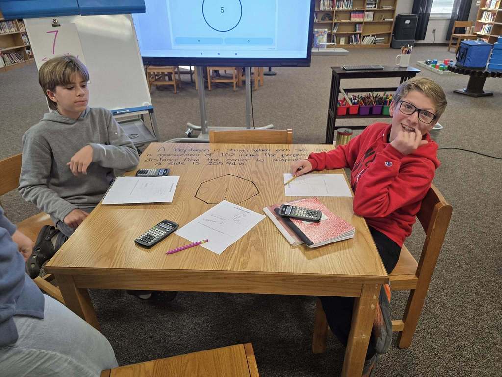 Students sit at a library table working together on a math activity, using calculators and paper while a digital display and whiteboard are visible in the background.