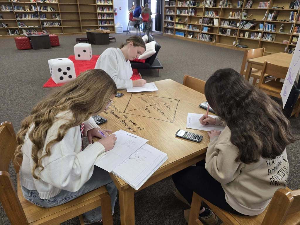 Students work independently and collaboratively at a library table, completing math assignments written on the tabletop with notebooks and calculators nearby.