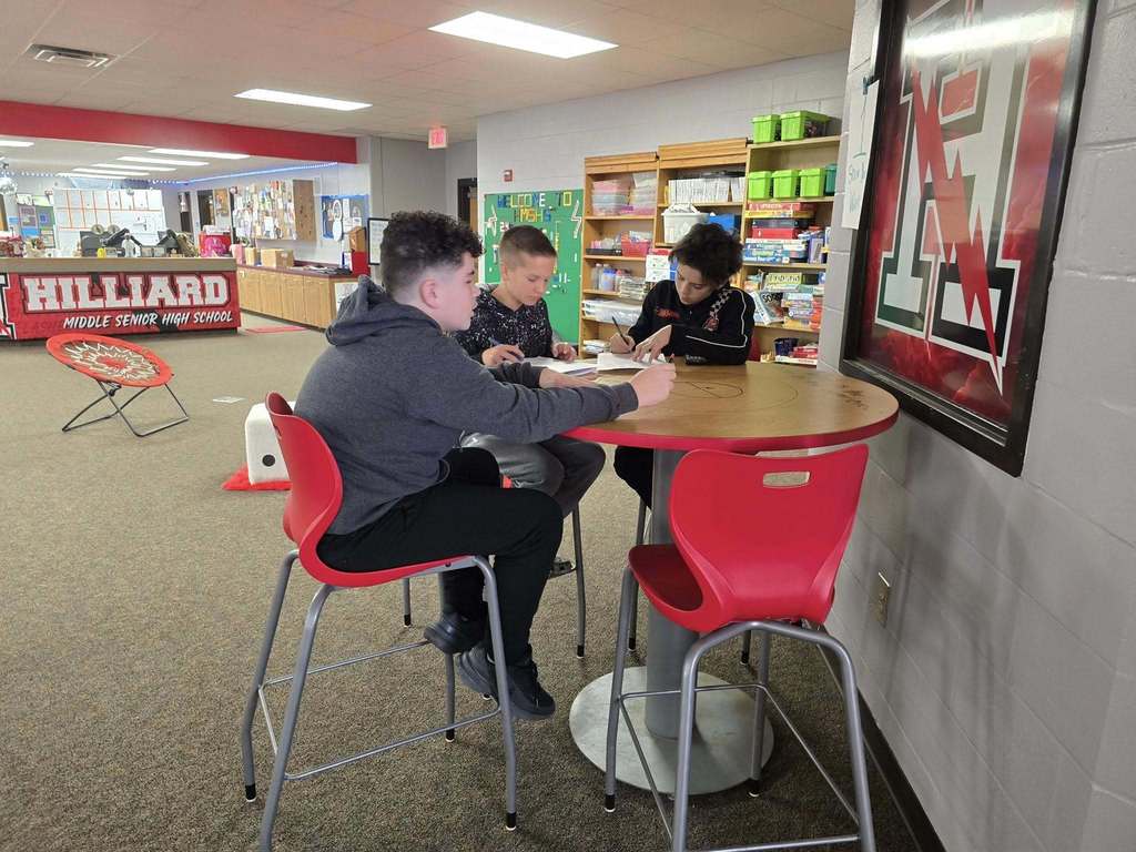Three students sit at a tall round table in a school library collaborating on written math problems, with bookshelves and school signage visible behind them.