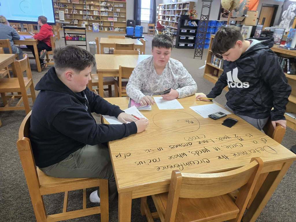 Students collaborate at a library table on a math task written on the tabletop, using pencils, notebooks, and calculators in a shared workspace.