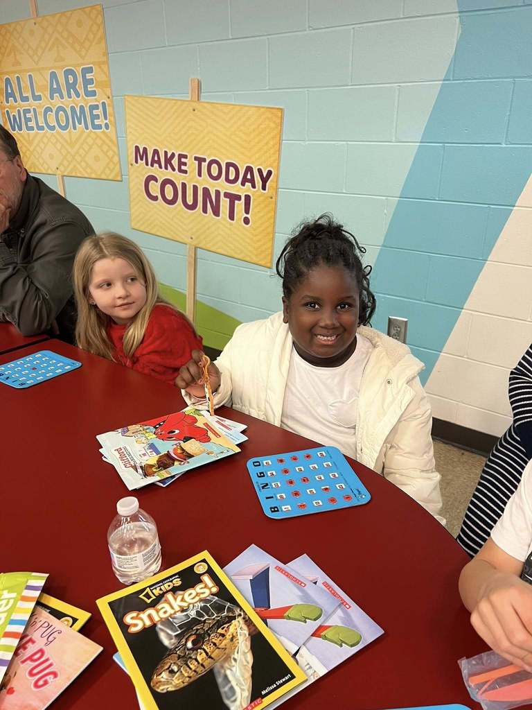 Two elementary students sit at a cafeteria table smiling while holding bingo cards and books, with posters reading “All Are Welcome” and “Make Today Count” on the wall behind them.