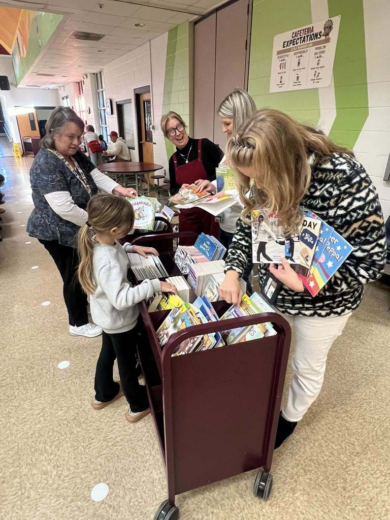 Staff members and volunteers stand behind rolling book carts while students browse and select books during the event.