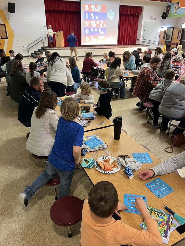 Students and adults sit closely together at cafeteria tables focused on bingo cards and books during the Bingo for Books event.
