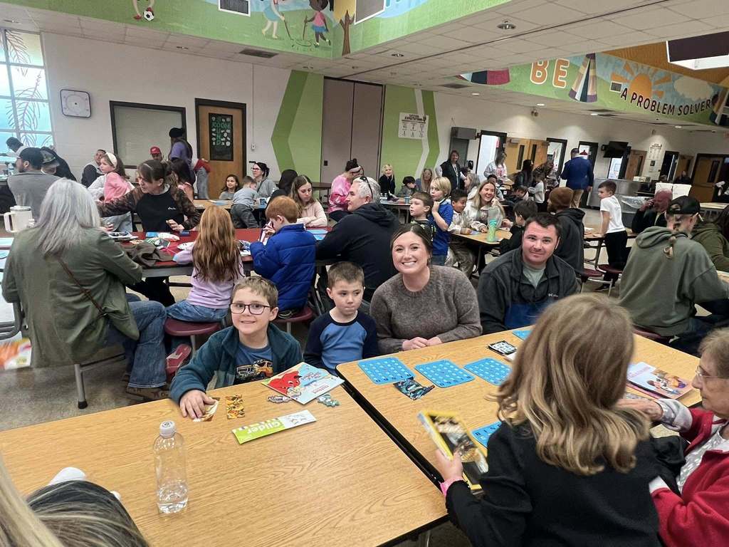 Families and students sit together at cafeteria tables playing bingo, with books, bingo cards, and reading materials spread across the tables.