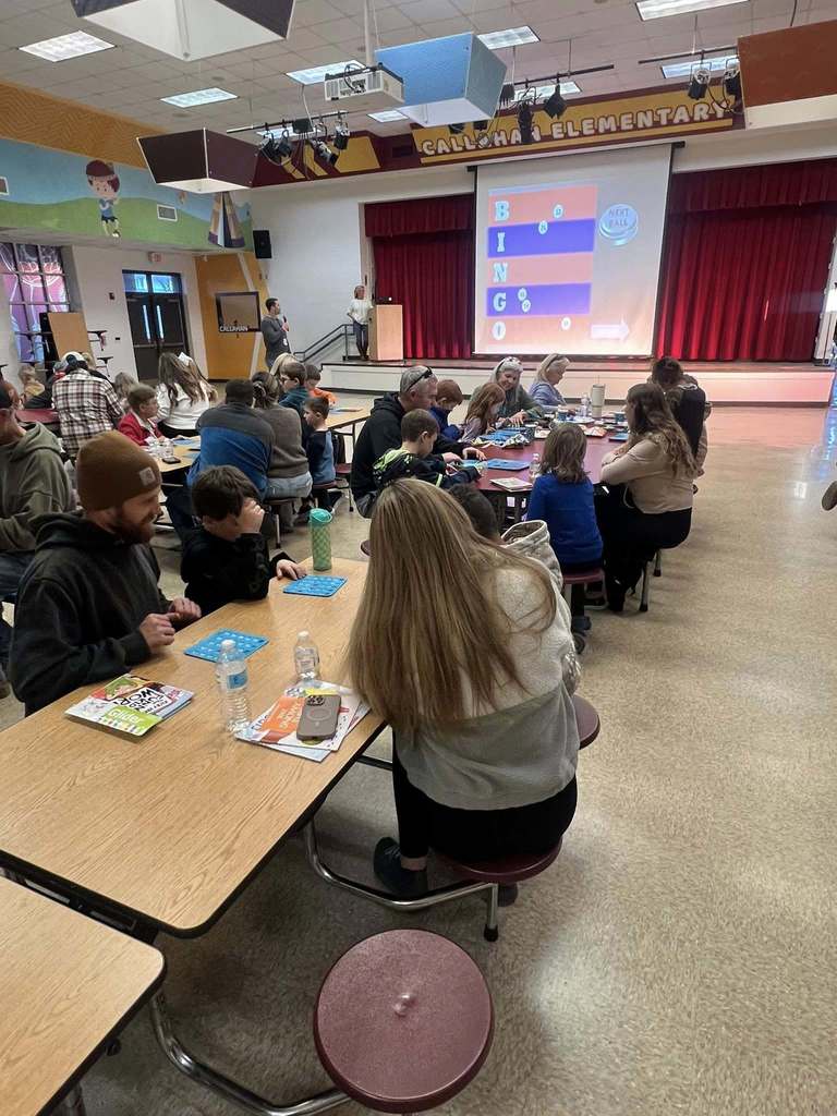 A wide view of the school cafeteria shows many families seated at tables playing bingo, with a large bingo board projected on a stage at the front of the room.