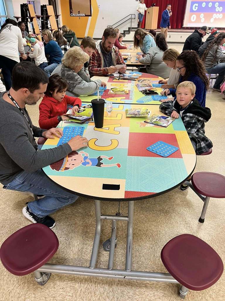 A group of students and adults sit around a round cafeteria table labeled “Callahan,” reviewing bingo cards and children’s books.