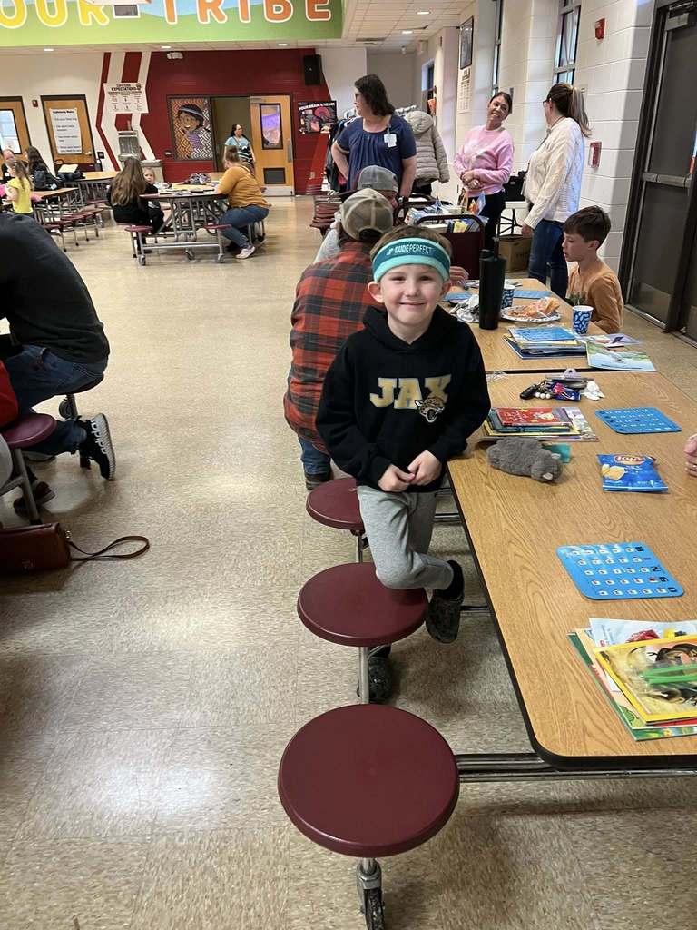 A young student stands smiling near a cafeteria table filled with books and bingo cards while families participate in the event in the background.