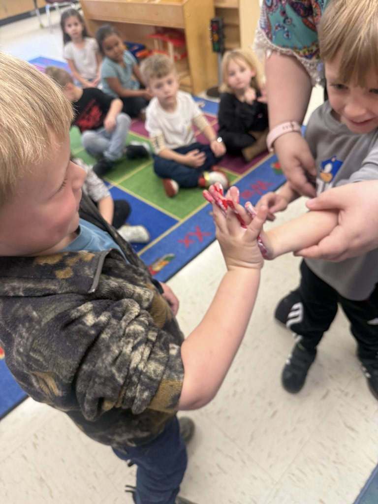 Two Pre-K students press their painted hands together while smiling, with a classroom chart labeled “Color Mixing” displayed behind them.