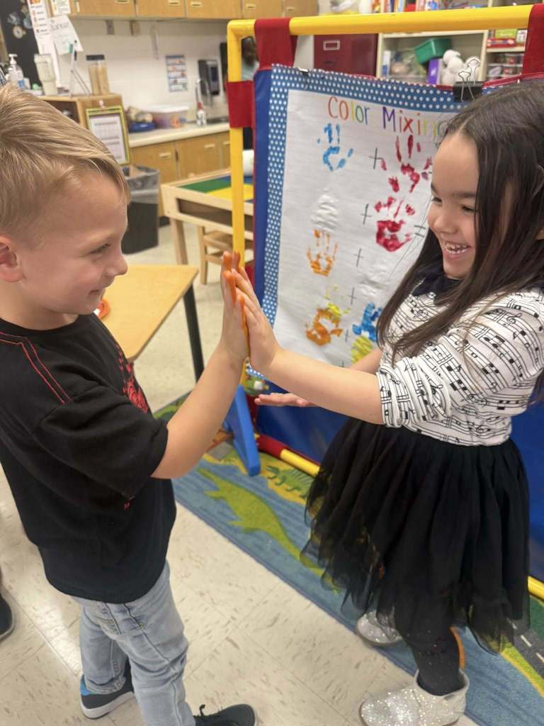 Two Pre-K students press their painted hands together while smiling, with a classroom chart labeled “Color Mixing” displayed behind them.